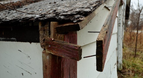 A view of a corner of a roof of an abandoned farmhouse. The roof is rotting and the boards along the edge of the roof have come unnailed. 