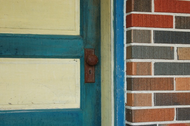 close up view of a door that is painted yellow and blue.  The door frame is also yellow and blue.  The door knob is rusted.  Some of the brick of the house is also in the photo