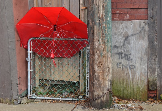 The red umbrella is wedged between the top of a chainlink gate and a wood door.