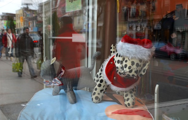 On display in a shop window are a stuffed toy cat and dog are dressed for Christmas.  THe dog is wearing a Santa hat.  Reflected in the window are people passing by carrying shopping bags. 