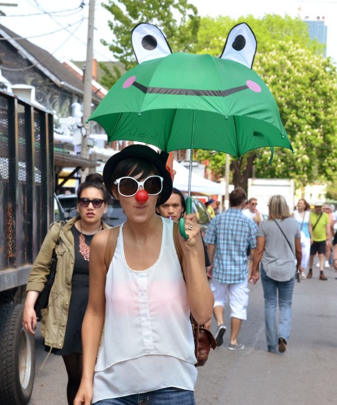 A woman has a large fake red nose and large white frames sunglasses.  She is carrying a green umbrella with a froggie face on it 