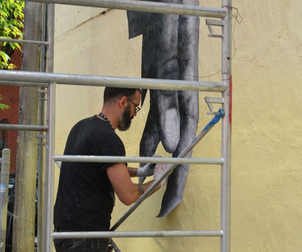 Street art of large hands that have been printed on paper and then pasted to a wall.  The artist is in the process of gluing a finger to the yellow wall. 