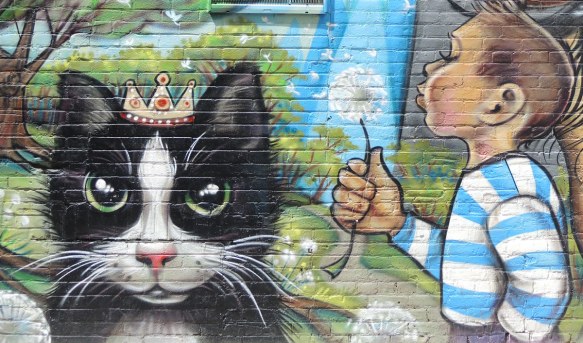 a large black and white cat is looking at the viewer. It is wearing a little crown. Beside the cat is a boy in a blue and white striped shirt who is blowing the seeds off a puffy white dandelion. 