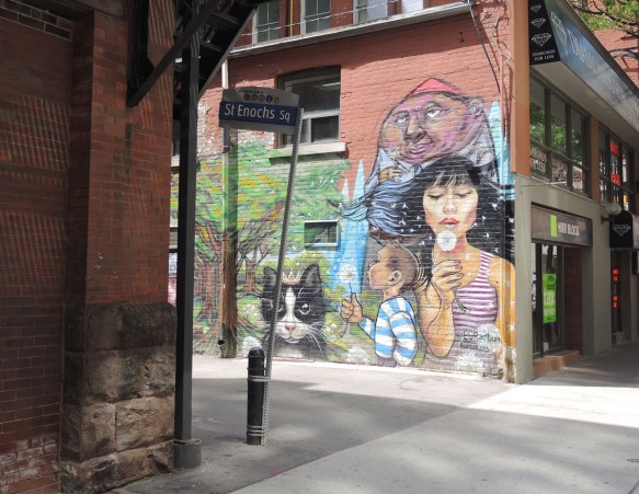 entrance to laneway, red brick building on the left (Massey Hall), mural painted on the wall on the right. Mural shows a woman & a boy blowing the seeds from white puffy dandelions. 