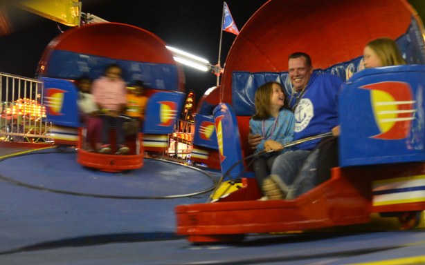 A father and his daughters are on a midway ride, spinning in red "seats" as they go around in circles. 