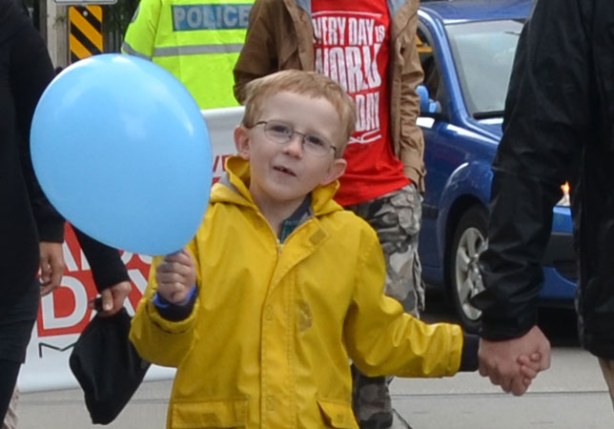 people at the AIDS walk in Toronto.  A boy in a yellow raincoat and holding a blue ballon is smiling for the camera. 