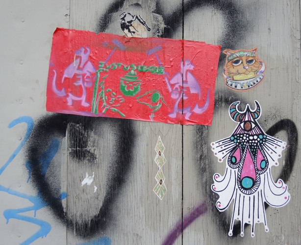 Graffiti in an alley,  a few stickers on a grey wood wall - the head of a cat, a small horse and an abstract drawing.