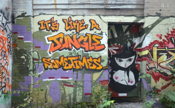 Large orange and yellow graffiti letters that say It's Like A Jungle Sometimes.  There is a door on the right side and on the door is a large black and white picture of a woman from the waist up.  She is bare chested with little pink hearts covering her nipples. 