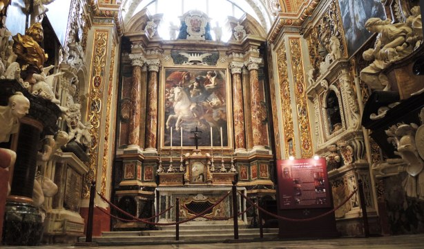 A chapel in a cathedral.  An altar with six very tall candles in front of a large painting of a man on a white horse.  The rest of the chapel is very ornately decorated. 