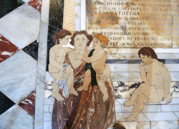 Mosaic tile work on the fllor - a woman holding two children while another woman sits on stone steps.  There are also words in the picture. 