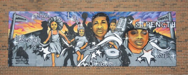 A large mural on a red brick building, painted to celebrate the United Nations Universal Declaration of HUman Rights.  This one includes the words strength, love, respect, unity.  It shows a 5 people engaged in various activities. 