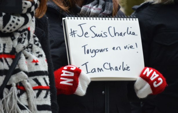 Close up of a sign that says 'Je suis Charlie Toujours en vie!  I am Charlie' in black letters on white paper.  The sign is being held by someone who is wearing red mitts with big CAN on them in white. 