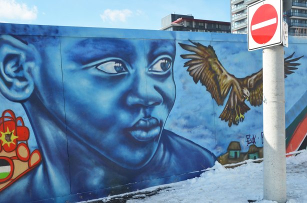 Colourful graffiti on hoardings around a construction site.  Large blue boy's face with a large bird taking flight beside him