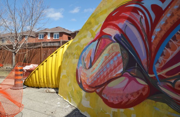 Close up of part of the mural of a skein of brightly coloured, multicoloured, wool on a bridge underpass, also the yellow part of a culvert that provides a passageway (tunnel) for the sidewalk.  Some houses are in the background. 