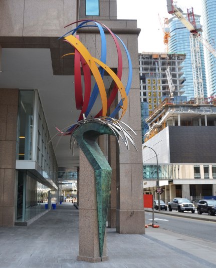 Public art on a corner called 'The Wave' by Ivan Kostov. curved pieces of coloured metal on top of a greenish pedestal. 