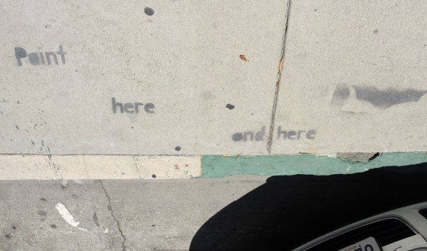 black paint with stencil graffiti on a sidewalk - words that say 'paint here and here' but there is a large space between each word 
