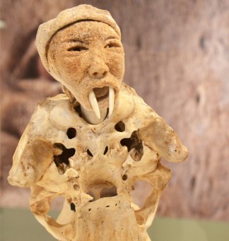 part of a sculpture by Manasie Akpaliapik made of bone, ivory and stone, a man like creature with a man's face but with walrus tusks