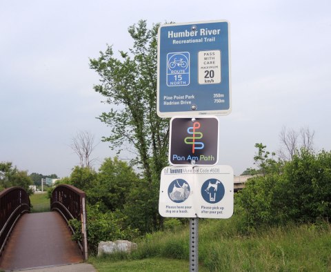 signs along the HUmber Recreational trail indicating the name of the trail, the cycle path number that it is, the fact that it is also the Pan Am Path, and lastly a sign that says dogs must be on a leash. 