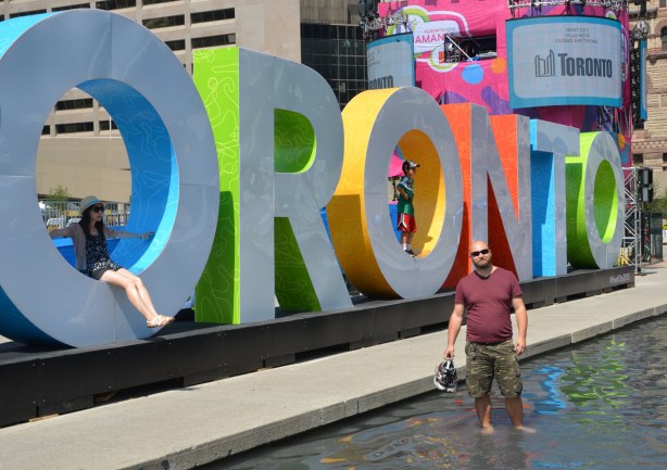 large three dimensional block capital letters that spell Toronto installed alongside the pool fountain in Nathan Phillips Square - a man stands in the water in front of the word Toronto 