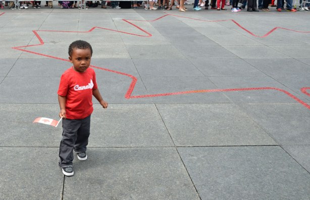 A young man stands in Dundas Square in an area that has been barricaded off and also in which a large maple leaf outline in red tape has been laid down.  He is wearing a red Canada T shirt and holding a small Canadian flag. 