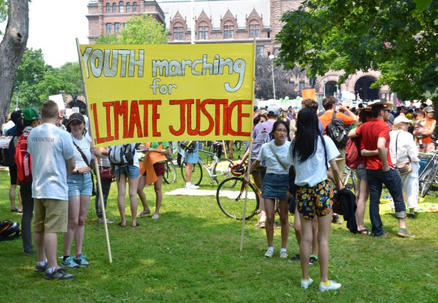 a groupd of young people standing beside a large yellow banner that says 'Youth marching for Climate Justice' at a protest rally in front of the Ontario parliament buildings. 