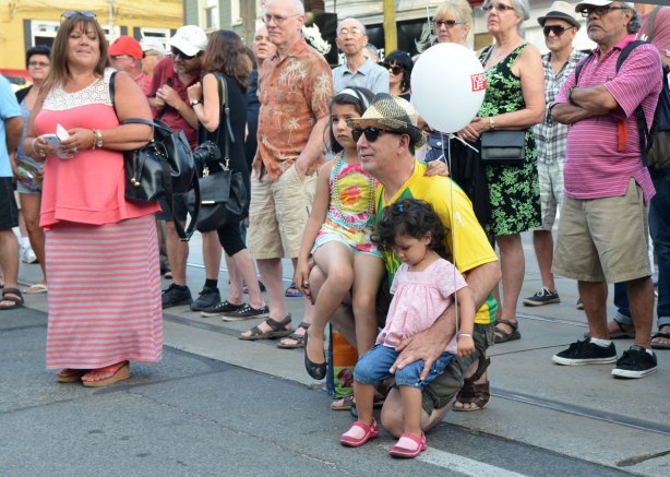 A man is squatting on the sidewalk with two girls on his knee.  Both girls have balloons.  They are part of a crowd watching a band perform in a street music festival 