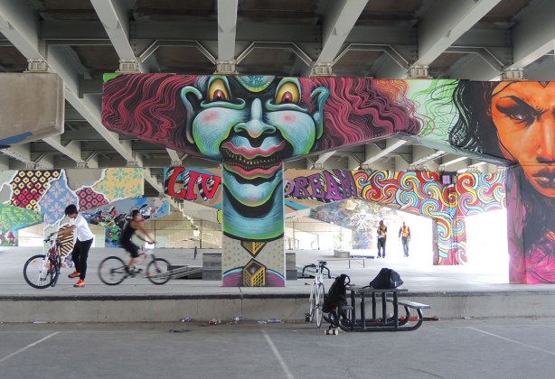 picture of art (mural) on a concrete support holding up a road above a skateboard and basketball park - guys on bikes in the park with many of the pillars in the photo including a large greyish blue clown face and an orange woman's face. 