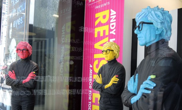statues of Andy Warhol, one pink, one yellow and one blue, in a window of an art gallery,  He's dressed in black, with black rimmed glasses and his hands are folded over his chest in all three statues.  Life sized. 