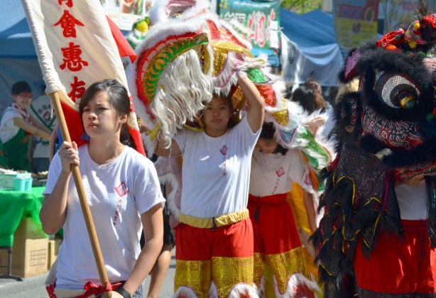 Two women under a white Chinese dragon costume and they are walking beside a black dragon, They are following a girl who is holding a white flag with red Chinese characters on it