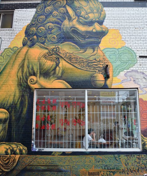 window of a Chinese restaurant with a young man sitting at a table by the window.  On the exterior wall surrounding that window is a painting of a large Chinese lion statue that is part of a larger mural 