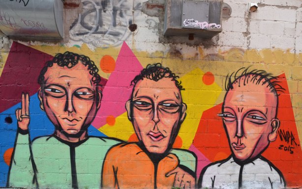 mural street art of three men from the waist up. All have short hair, one with green shirt, one with orange shirt and the third with a white shirt, 