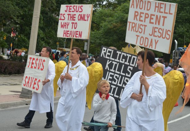 some people wearing white robes and large gold angel wings are walking in a parade. Three of them are holding signs. The first signs says Jesus Saves. The second sign says Jesus is the only way to heaven. The third sign says Avoid hell repent trust Jesus today. 