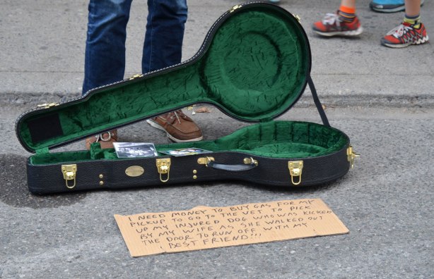 a street musician stands beside his empty banjo case. In front of the case is a note that says " I need money to buy gas for my pickup to go to the vet to pick up my injured dog who was kicked by my wife as whe walked out the door to run off with my best friend"