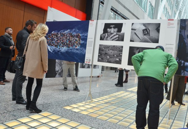 Three people are looking at a series of photographs on display. One of the photos is a boat carrying refugees, taken from above, the boat is packed full 