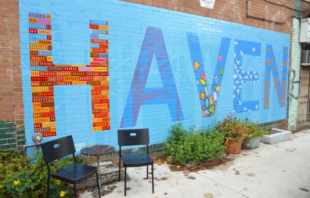 two chairs and a small table in front of a wall with a mural on it. The mural is the word Haven. Each letter is decorated in a different way. 