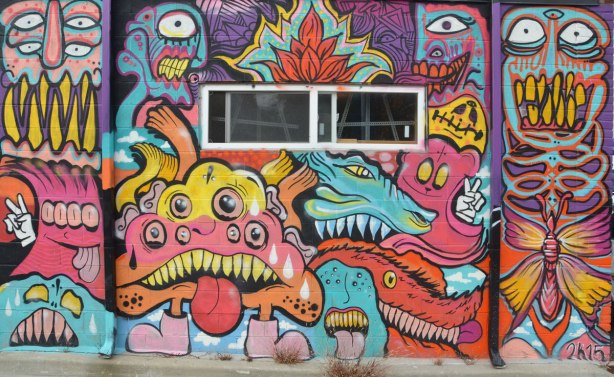 mural on a wall, with a window in the middle, many blob creatures with different number of eyes in blues, reds and purples, many with tongues sticking out, one giving a peace sign with white hand