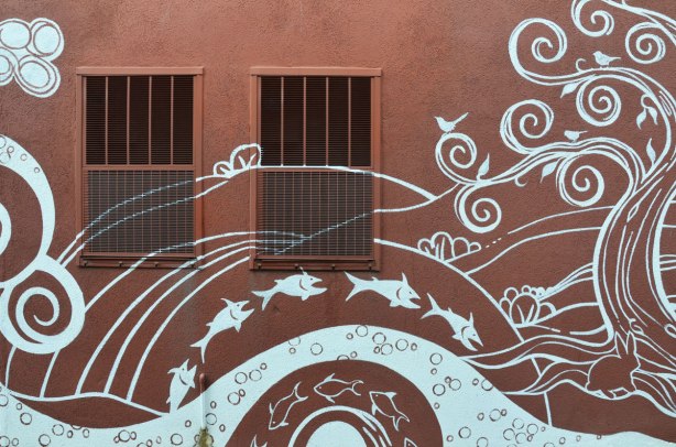 part of a mural by Monica on the Moon, white line drawings on brown background, across the back of a building in a laneway - jumping fish, a tree