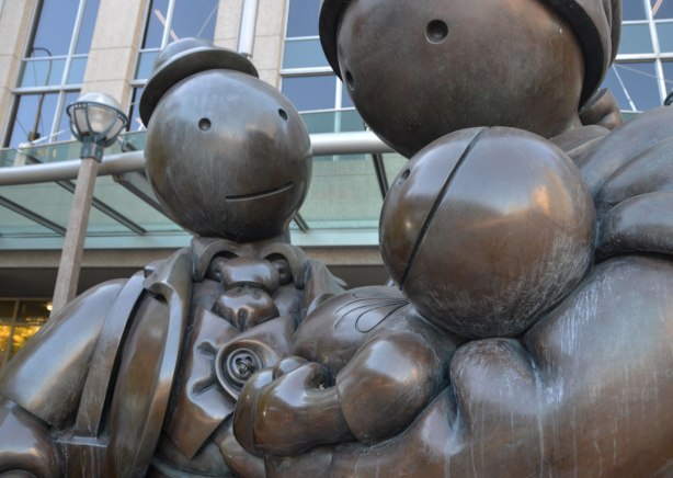 close up of a sculpture by Tom Otterness of a family of three, mother, father and baby in arms. Father is carrying a suitcase. Title of sculpture is Immigrant Family