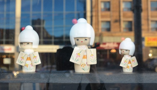 three little Japanese wooden dolls with white hair and white kimonos standing inside a window. Reflections of the stores across the street are behind them. 