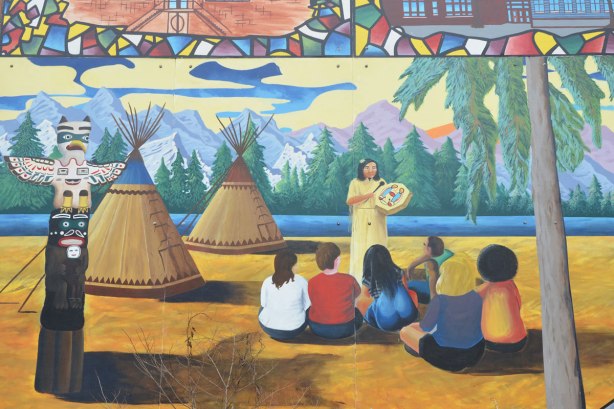 a group of an indigenous peoples are sitting and listening to a woman who is holding a drum. two teepees are in the picture as well