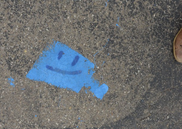 A blue square shaped painted spot on the pavement. Two eyes and a smiling mouth have been drawn on top in darker blue
