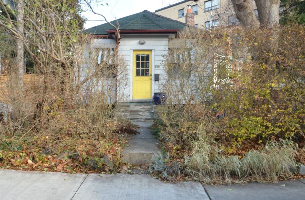 little narrow white bungalow with a yellow front door set back from the road. There is a straight walkway from the sidewalk to the door, and there is a lot of shrubery in the front yard especially near the sidewalk. 