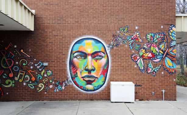 A brightly coloured mural on a brick wall. A large oval face with music symbols on the left and butterflies on the right. Called united freedom and painted by essencia. 