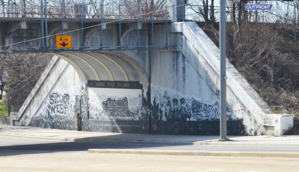 mural in black, white, and grey about the first railway in Ontario, 1853, that was built here, and where the railway still runs as the GO line to Aurora - a photo of the mural from across the street, with its three parts, the words St. Clair West on the left, the locomotive in the middle and the railway workers on the right