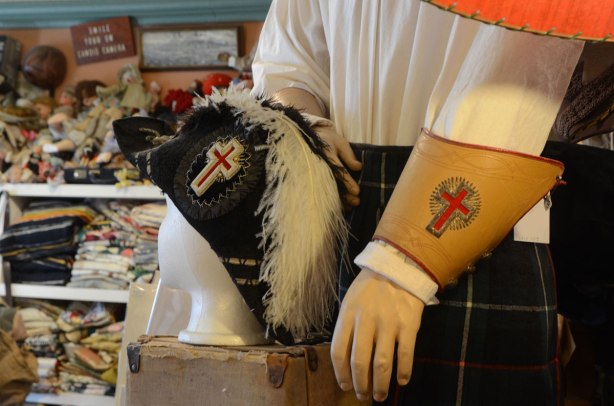 An old black military hat with a white feather in it sits on a head mannequin. Beside it is another mannequin wearing a tartan kilt and a white shirt. The shirt has a brownish leather cuff with a red cross on it. 