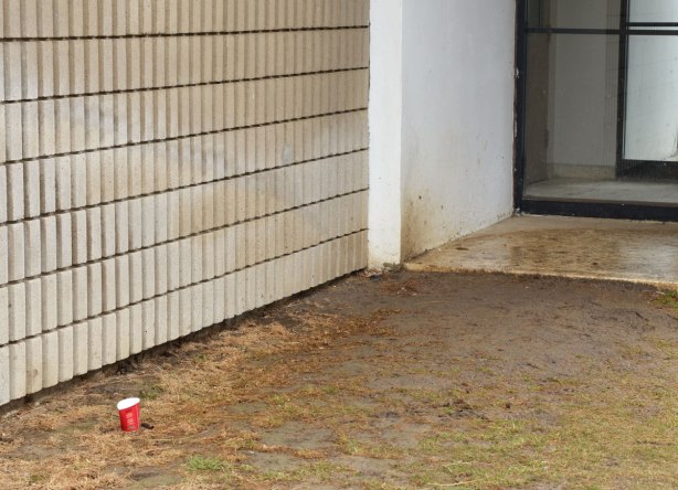 a small red empty Tim Hortons coffee cup sits on the muddy ground outside the back entrance of an older grey brick apartment building