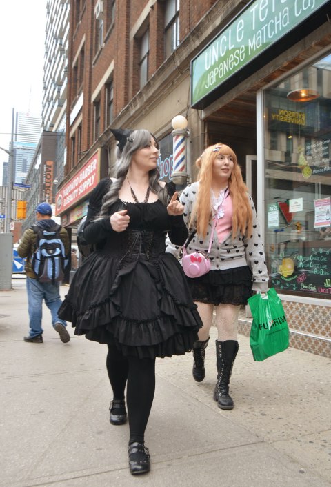 two women dressed up and walking down the street. One is in a puffy black dress, black tights and black shoes. The other woman has long red hair and a polka dot sweater on. 