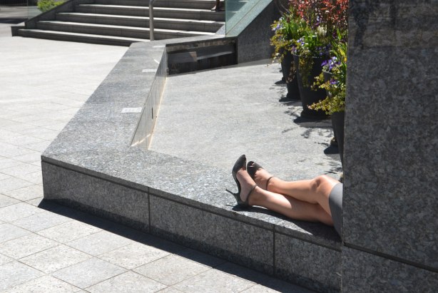 A woman in black high heel shoes is sitting on a low stone wall outside. You can only see her from the thigh down. He legs are crossed at the ankle. 
