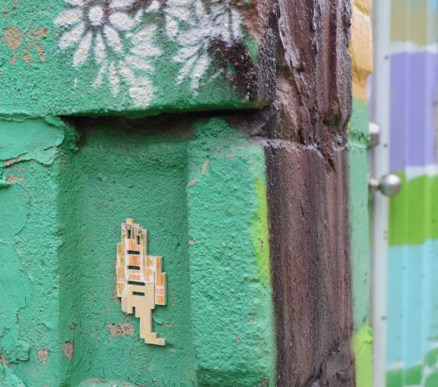a little wooden stikman with only one leg on a green concrete block wall, with white stencilled daisies above him, street art in an alley