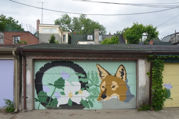 garage door in an alley painted with a mural of a fox head beside a bicycle wheel with trilliums growing up in front of it. 